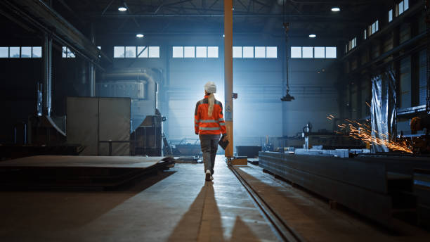 istock Professional Heavy Industry Engineer/Worker Wearing Safety Uniform and Hard Hat Uses Tablet Computer. Serious Successful Female Industrial Specialist Walking in a Metal Manufacture Warehouse. 1290244494
