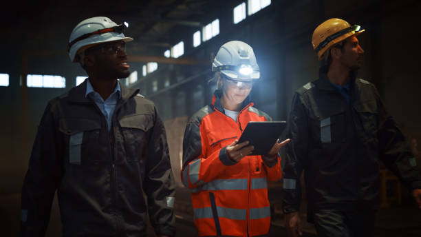 tre diversi ingegneri e lavoratori multiculturali dell'industria pesante in uniform walk nella fabbrica di acciaio scuro che usano torce sui loro cappelli duri. female industrial contractor utilizza un tablet. - data mining foto e immagini stock