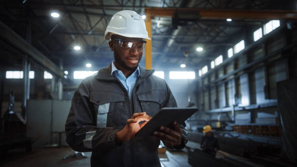 istock Professional Heavy Industry Engineer Worker Wearing Safety Uniform and Hard Hat Uses Tablet Computer. Smiling African American Industrial Specialist Walking in a Metal Construction Manufacture. 1290244537