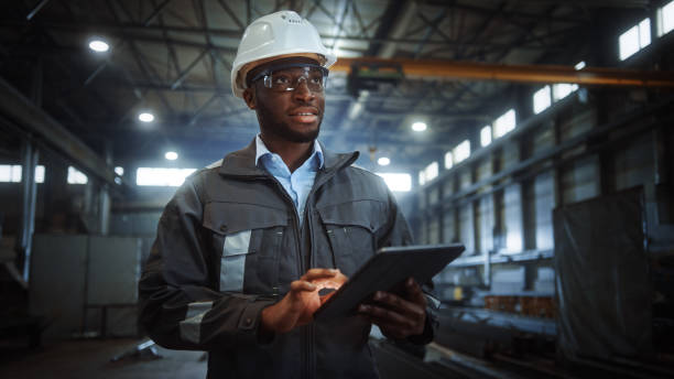 istock Professional Heavy Industry Engineer/Worker Wearing Safety Uniform and Hard Hat Uses Tablet Computer. Smiling African American Industrial Specialist Walking in a Metal Construction Manufacture. 1290244541