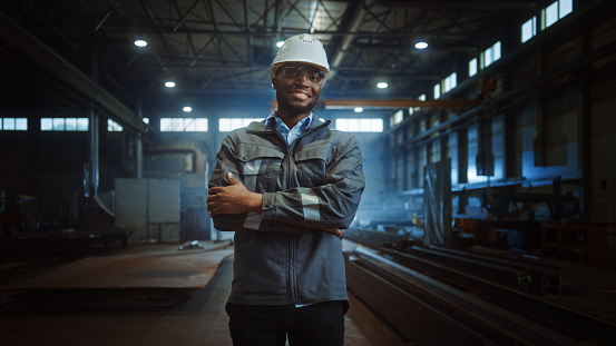 Happy Professional Heavy Industry Engineer/Worker Wearing Uniform, Glasses and Hard Hat in a Steel Factory. Smiling African American Industrial Specialist Standing in a Metal Construction Manufacture.