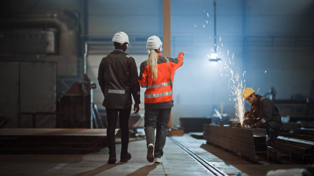 istock Two Heavy Industry Engineers Walk Away from Camera in Steel Factory and Discuss Work. Industrial Worker Uses Angle Grinder in the Background. African American Specialist Talks to Female Technician. 1290244577