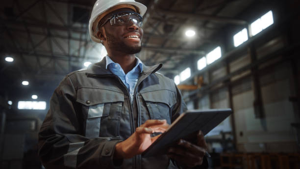 istock Professional Heavy Industry Engineer/Worker Wearing Safety Uniform and Hard Hat Uses Tablet Computer. Smiling African American Industrial Specialist Walking in a Metal Construction Manufacture. 1290244595