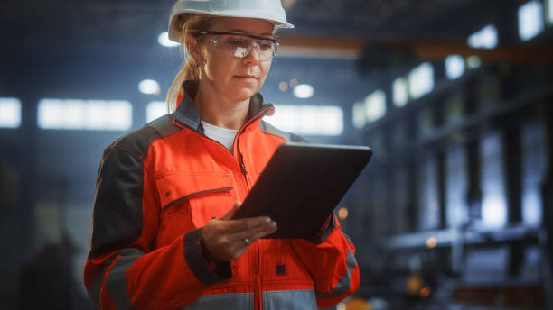 istock Professional Heavy Industry Engineer/Worker Wearing Safety Uniform and Hard Hat Uses Tablet Computer. Serious Successful Female Industrial Specialist Standing in a Metal Construction Manufacture. 1290244612