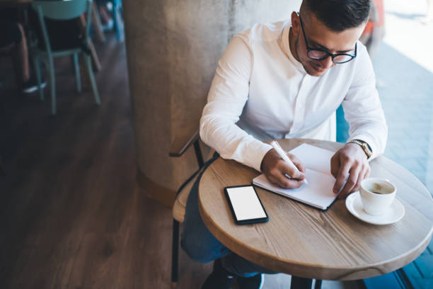 istock Focused man writing in notebook while sitting at table with smartphone and coffee 1290256869