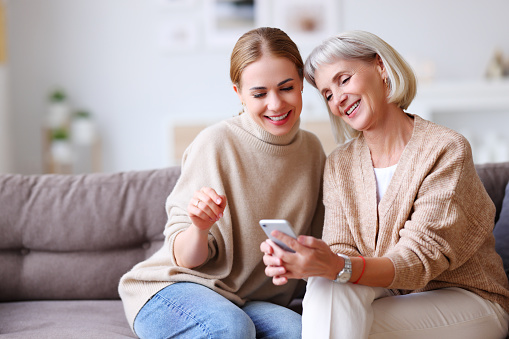 Delighted adult and aged women smiling and browsing social media on smartphone while resting on couch at home together