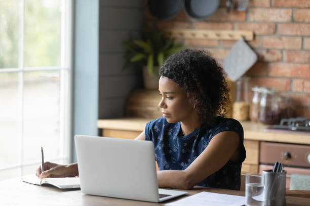 Focused young african american woman writing notes, working on computer.