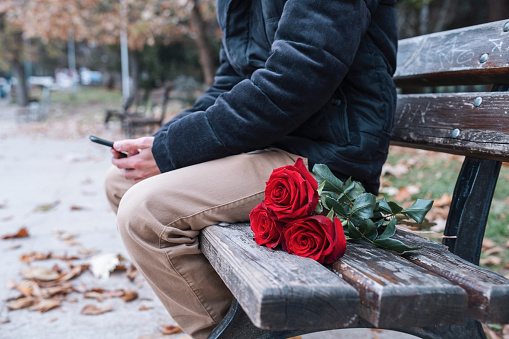 Unrecognizable man sitting on bench in public park and waiting for his girlfriend with bouquet of red roses.