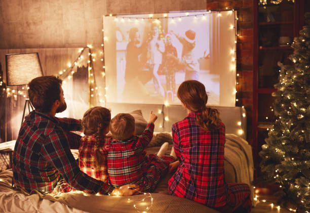 family mother father and children watching projector, film, movi happy family in checkered pajamas: mother father and children watching projector, film, movies with popcorn in christmas holiday evening   at home merry christmas family stock pictures, royalty-free photos & images