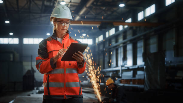 istock Professional Heavy Industry Engineer/Worker Wearing Safety Uniform and Hard Hat Uses Tablet Computer. Serious Successful Female Industrial Specialist Standing in a Metal Construction Manufacture. 1290267807