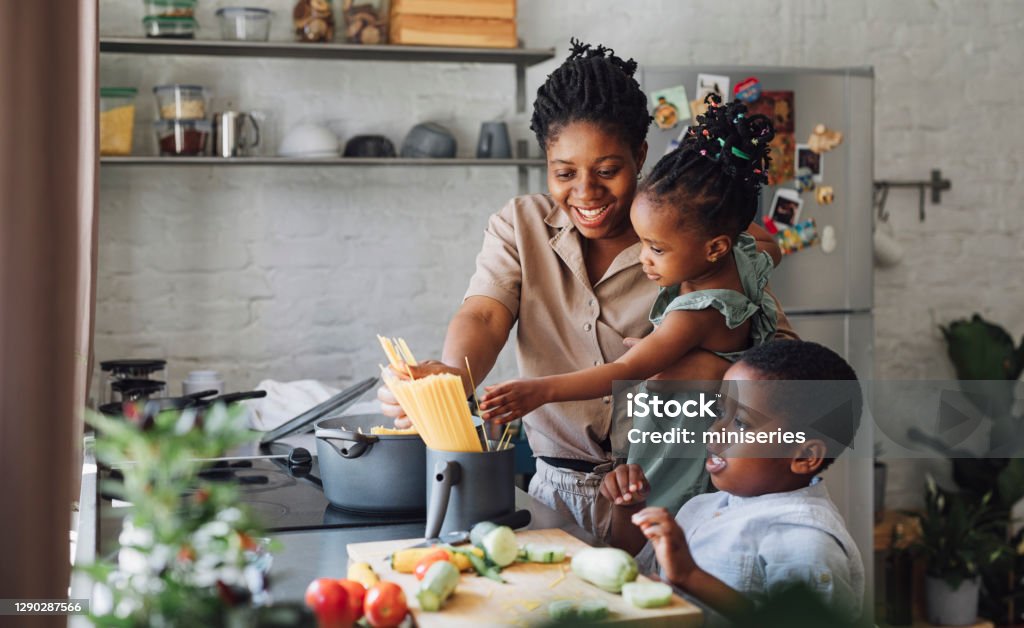 Mother, Daughter and Son Preparing Spaghetti and Vegetables for Lunch over a Cutting Board Happy African American mother, daughter and son preparing spaghetti and vegetables for lunch over a kitchen sink. Family Stock Photo