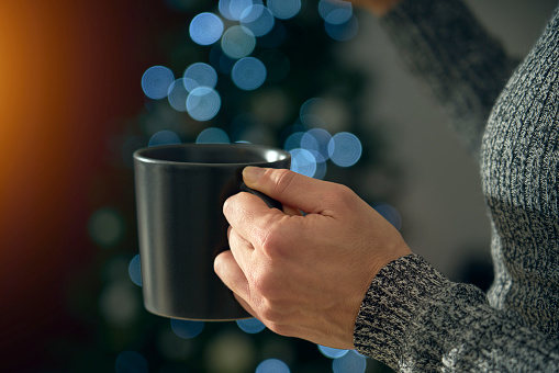A young woman drinks a cup of tea or hot chocolate to warm up from the cold of winter. In the background the Christmas tree lights create a bokeh effect to celebrate the holy festivities.