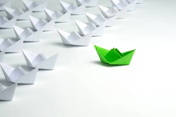 Leadership concept with Green paper ship standing out from the group of white ships on white background.