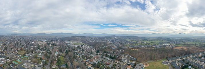 Wide-angle of peaceful small town under cloudy sky and with mountains as background. Aerial view of slightly cloudy blue skyline above the suburbs. Rural town landscape