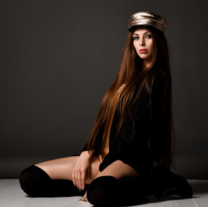 Young pretty sexy woman model with long straight hair in black stylish elegant clothes, cap and high socks sitting over dark grey background. Fashion, stylish look for women, sexy elegant wear concept
