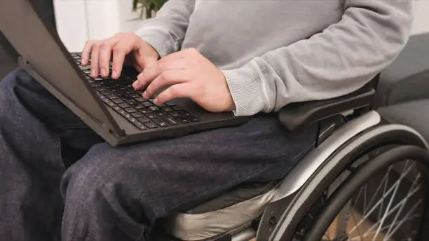Young man in wheelchair with laptop working remotely from home, using internet and looking at screen.