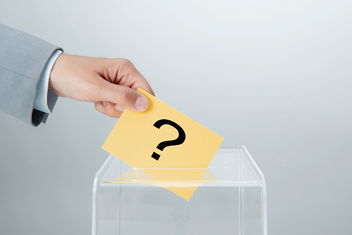Human hand of a businessperson is inserting yellow envelope  with black big question mark into ballot box in front of gray background.