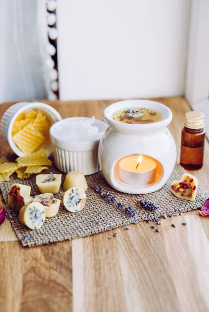 istock Using homemade mini wax melts in aromatherapy lamp diffuser at home interior concept. Melts making ingredients on table for unbleached beeswax, solid coconut oil, essential oil, dried flowers. 1290337363