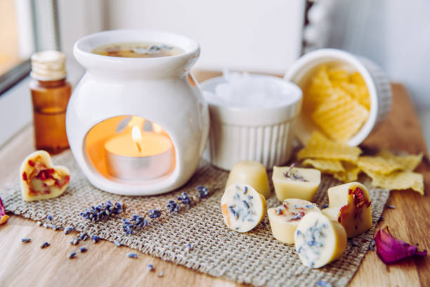 istock Using homemade mini wax melts in aromatherapy lamp diffuser at home interior concept. Melts making ingredients on table for unbleached beeswax, solid coconut oil, essential oil, dried flowers. 1290337367