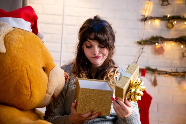 Stylish happy girl opening Christmas gift box sitting on a giant bear. Girl opens Christmas gift. Woman and a giant teddy bear home alone for Christmas.