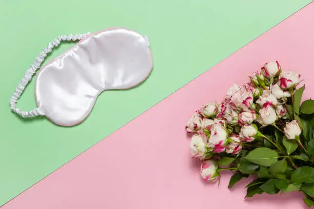Pink silk sleep mask, bouquet of small roses on pastel two-color background pink and green, copy space, flat lay. March 8, February 14, St. Valentine's, Mother's, Women's day celebration concept.