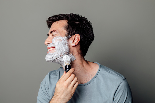 A smiling Caucasian man applying shaving cream with a shaving brush in the morning.