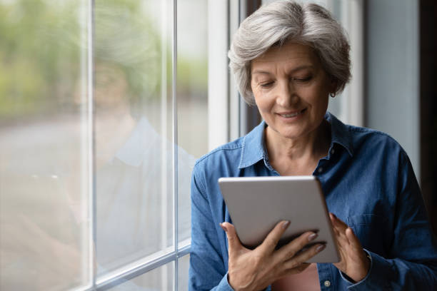istock Close up smiling mature woman using tablet, standing near window 1290347027