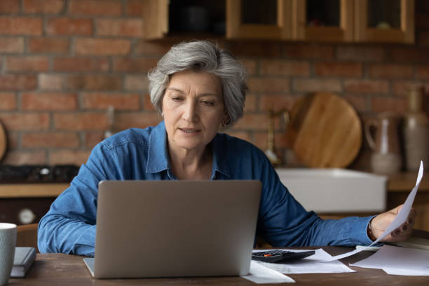 istock Smiling mature woman using laptop, checking financial documents at home 1290347065