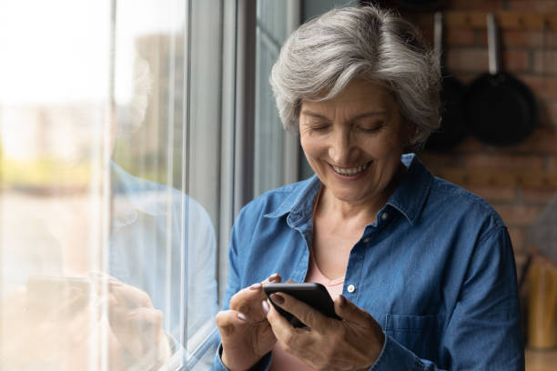 istock Close up smiling mature woman using smartphone, standing near window 1290347078