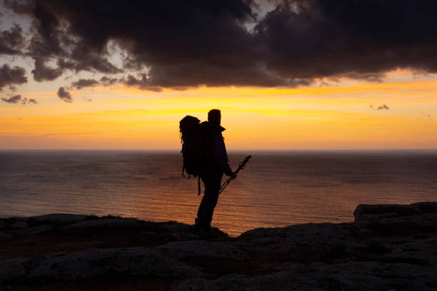 istock Man with backpack walking alone near the sea at sunset 1290351703