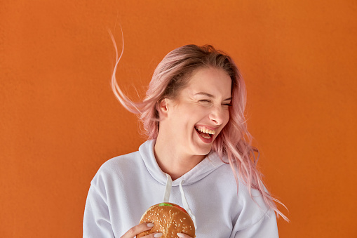 A happy woman holds a large hamburger in her hand and laughs. A woman on a diet. The concept of harmful nutrition. Orange background.