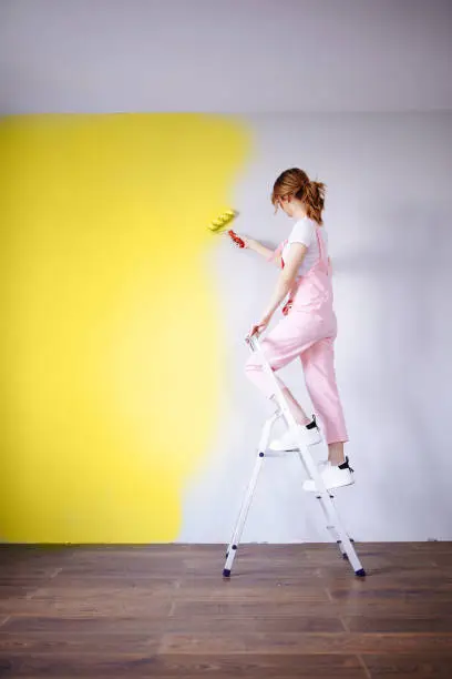 A woman in a pink jumpsuit is painting a wall. Renovation and new life for old things. Brushes and roller. Self-repair. Professional help.