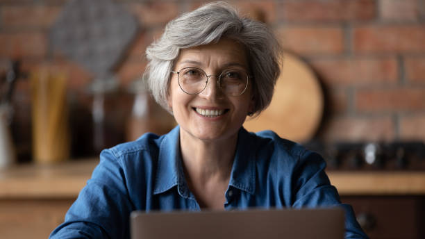 hoofd ontsproten portret dat rijpe vrouw glimlacht die glazen draagt gebruikend laptop - gesprek coaching detail stockfoto's en -beelden