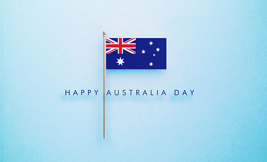 Tiny Australian flag sitting next to Happy Australia Day Message on light blue background. Horizontal composition with copy space. Directly above. Australia Day concept.