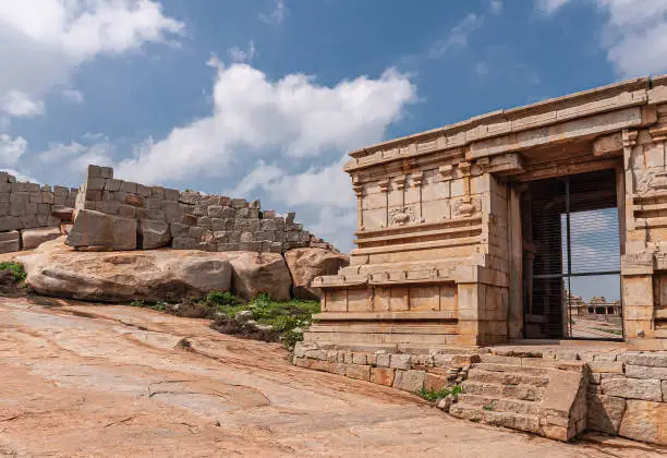 Hampi, Karnataka, India - November 4, 2013: Ruinous Kadelekalu Ganesha temple. Side brown stone buildings with screen and look-through. Gray stone wall and blue cloudscape.