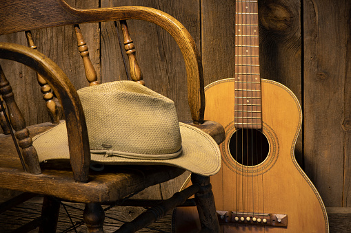 A cropped saloon chair with a cowboy hat on the seat. A vintage parlor guitar leans against a barnwood wall. A partial holiday wreath with illumination edges the top of the frame, Copy space available