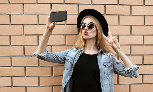 Pretty woman taking selfie picture by smartphone blowing kiss wearing a black round hat over brick wall background
