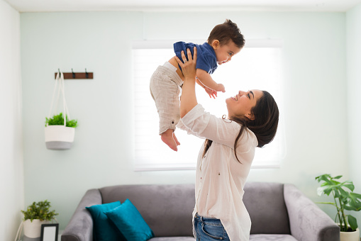 Happy young woman holding up her baby boy up while standing in living room