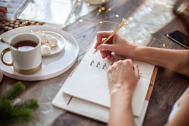 Young woman writing plans or goals for New Year 2021 while her daughter crafting Christmas trees Young woman drinking tea and writing plans or goals for New Year 2021 while her daughter crafting Christmas trees from paper cone, yarns and buttons with placed stars and fairy lights on wooden table. arranging stock pictures, royalty-free photos & images