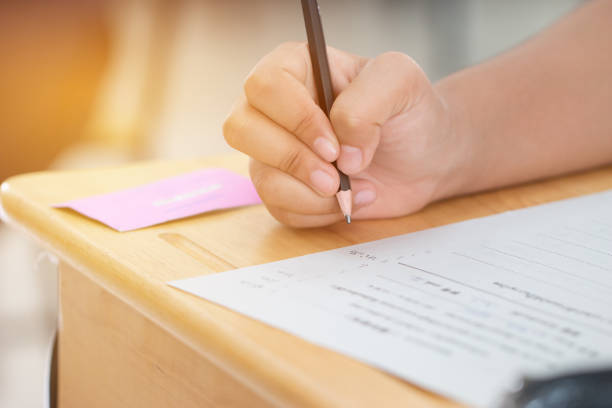 istock Education test concept : Hands student holding pen for testing exams writing answer sheet or exercise for taking fill in exam carbon paper computer on wood table at classroom in university. 1290423437