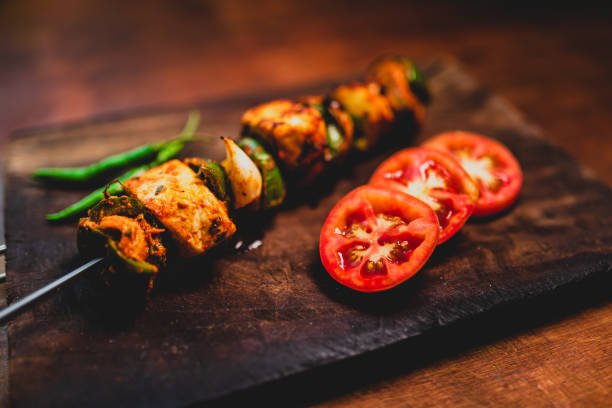 Paneer tikka masala flame frilled on skewer. Close up of paneer tikka on a skewer with fresh tomatoes, green chilli and vegetables. Spicy Indian starter food on a dark wooden plate. char grilled photos stock pictures, royalty-free photos & images