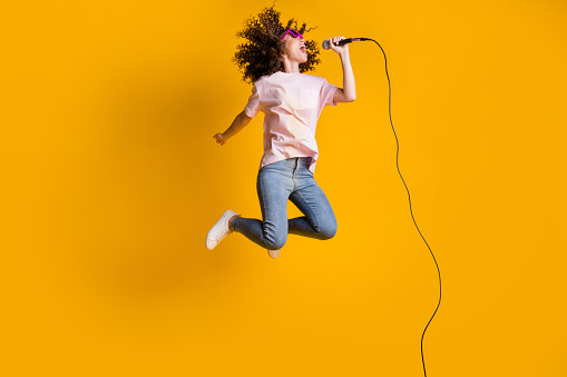 Photo portrait full body view of ecstatic woman jumping up singing into microphone isolated on vivid yellow colored background.