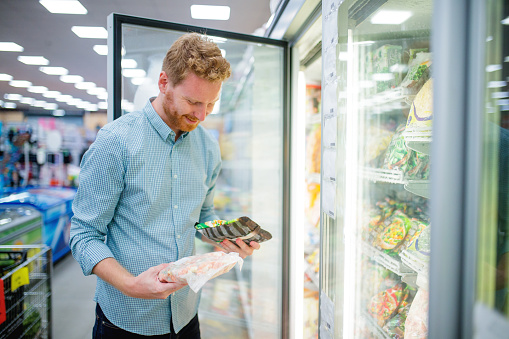 Cheerful young man in supermarket reading nutrion label on frozen food, standing in front of open freezer, copy space