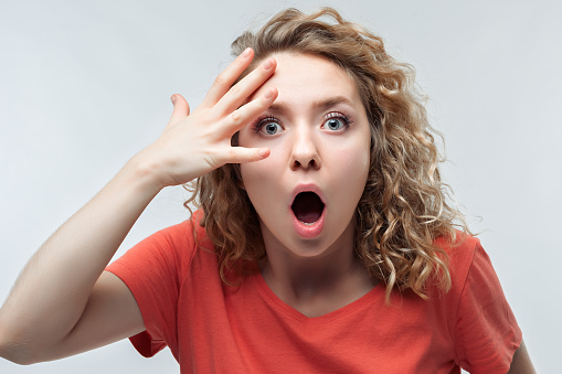 Young shocked woman with curly hair, dressed in casual t-shirt expressing surprise on camera. Human emotions, facial expression concept. Studio shot, white background