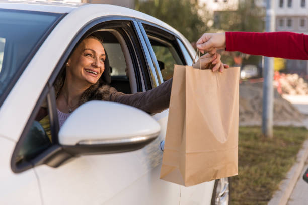Cheerful mature woman receiving her curbside pickup order Cheerful mature woman sitting in driver's seat in her car and receiving a paper bag with her takeaway order from unrecognizable saleswoman on a curbside pickup. curbsidepickup stock pictures, royalty-free photos & images