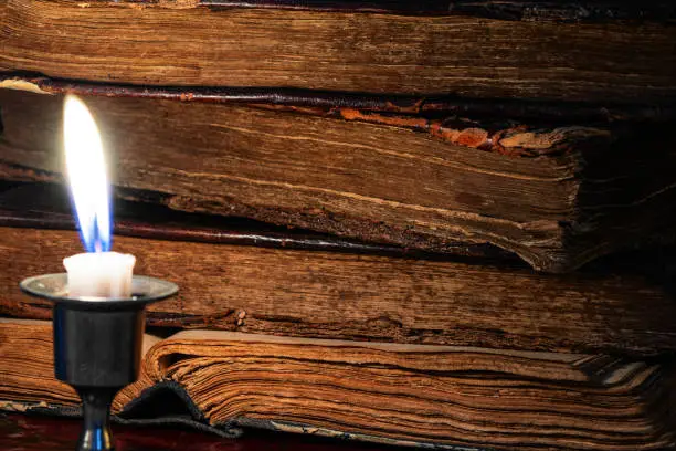 Stack of old worn leather-bound books and burning candle in the dark. Selective focus. Low key