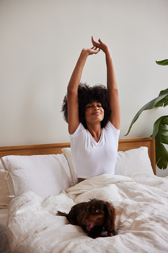 Young African American woman stretching and smiling while waking up in bed with her dog in the morning