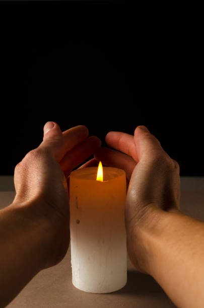 istock Vertical image.Warm candle flame, white cylindrical candle and human hands against drk background.Empty space 1290475792