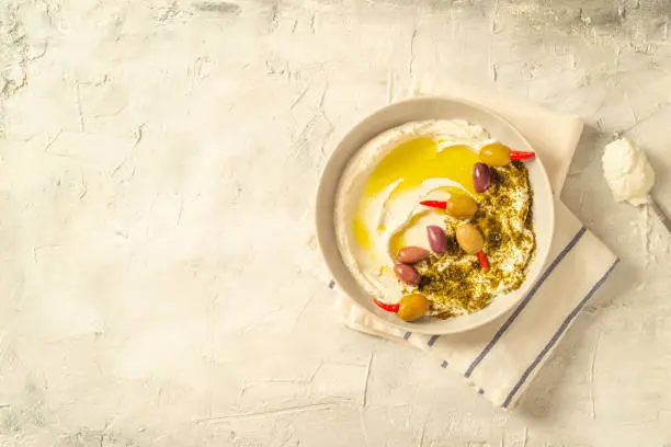 Photo of Popular middle eastern appetizer labneh or labaneh, soft white goat milk cheese with olive oil, hyssop or zaatar, olives on grey table, top view ,flat lay with copy space.