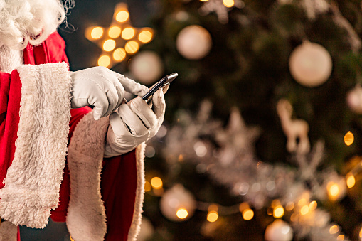 Santa Claus sitting in sofa next to Christmas tree and checking e-mails on smartphone.